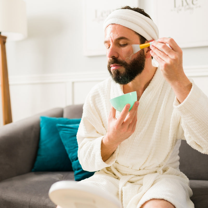 Man in a robe applying cream to his face with a brush, sitting on a couch.