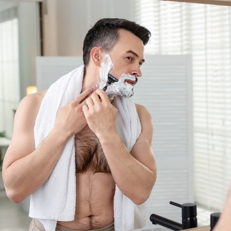 Man shaving his beard with a towel around his neck in a bathroom.