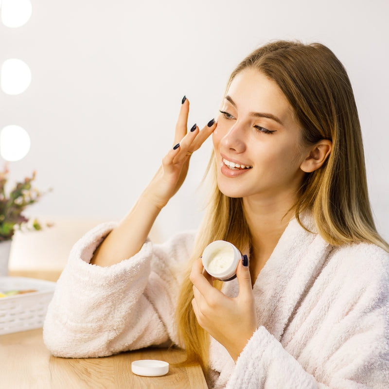 Woman in a robe applying cream to her face in a bright room.