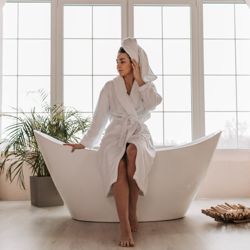 Woman in a white robe sitting on a modern bathtub with a towel wrapped around her head, in a bright bathroom.